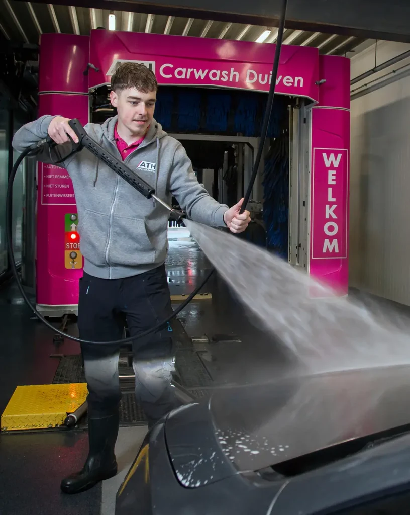 Een man spuit water op een auto in een moderne carwash met roze accenten.